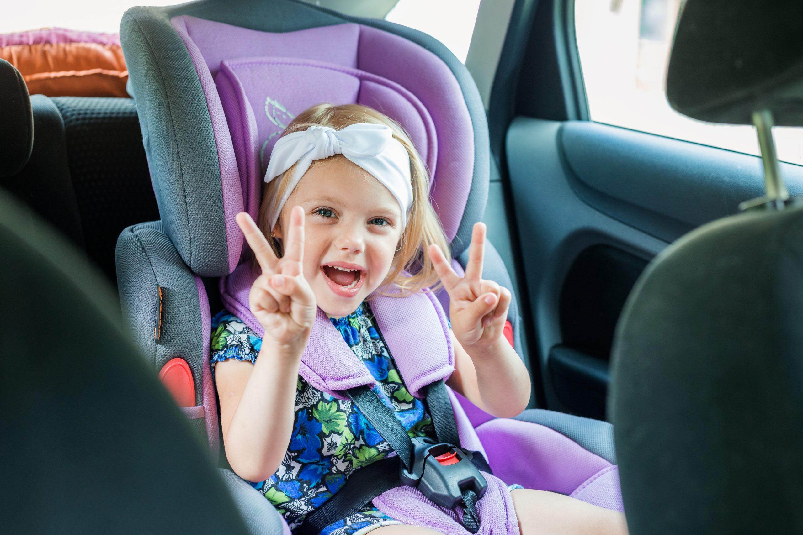 Nasmejana štiriletna deklica sedi v avtosedežu, pripeta z varnostnim pasom se veseli vožnje.