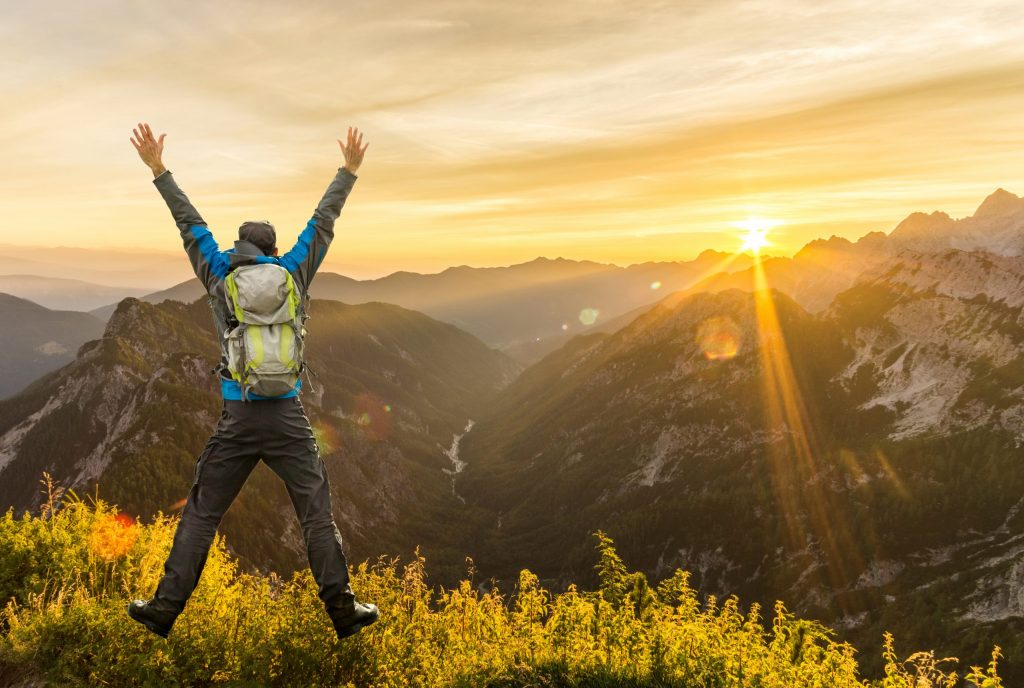 Veseli hribolazec z rugzakom skače z iztegnjenimi rokami na razgledni točki med gorami.